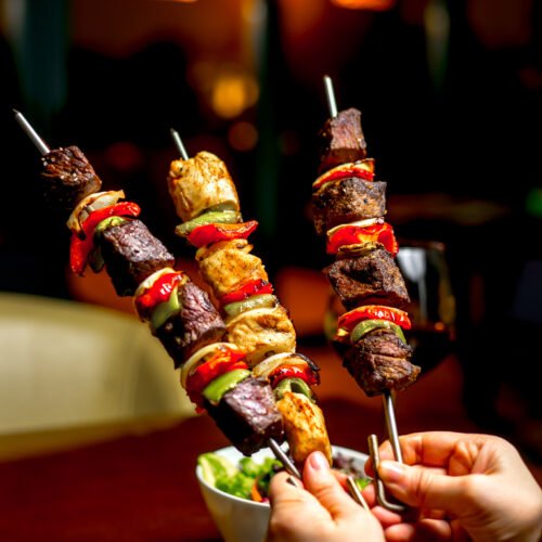 side view of hands holding shish kebab with colorful bell peppers and fresh vegetable salad on a wooden table